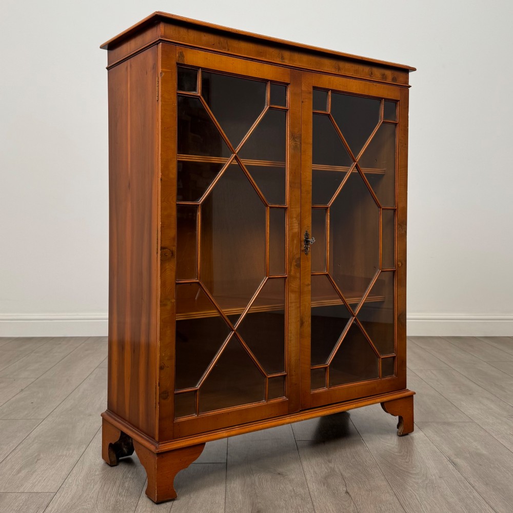 An antique yew wood display cabinet featuring two astragal glazed doors with geometric glazing bars, housing books on internal shelves, and resting on bracket feet. View X