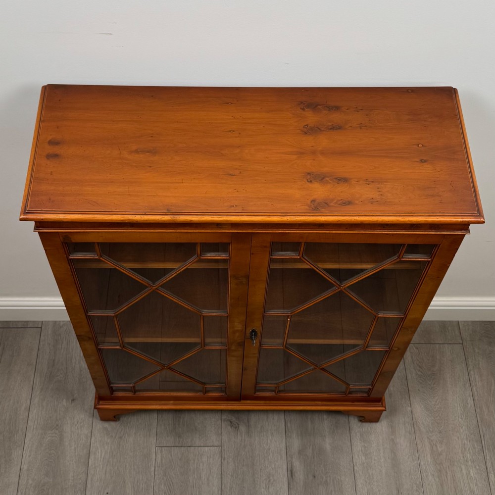 An antique yew wood display cabinet featuring two astragal glazed doors with geometric glazing bars, housing books on internal shelves, and resting on bracket feet. View XI