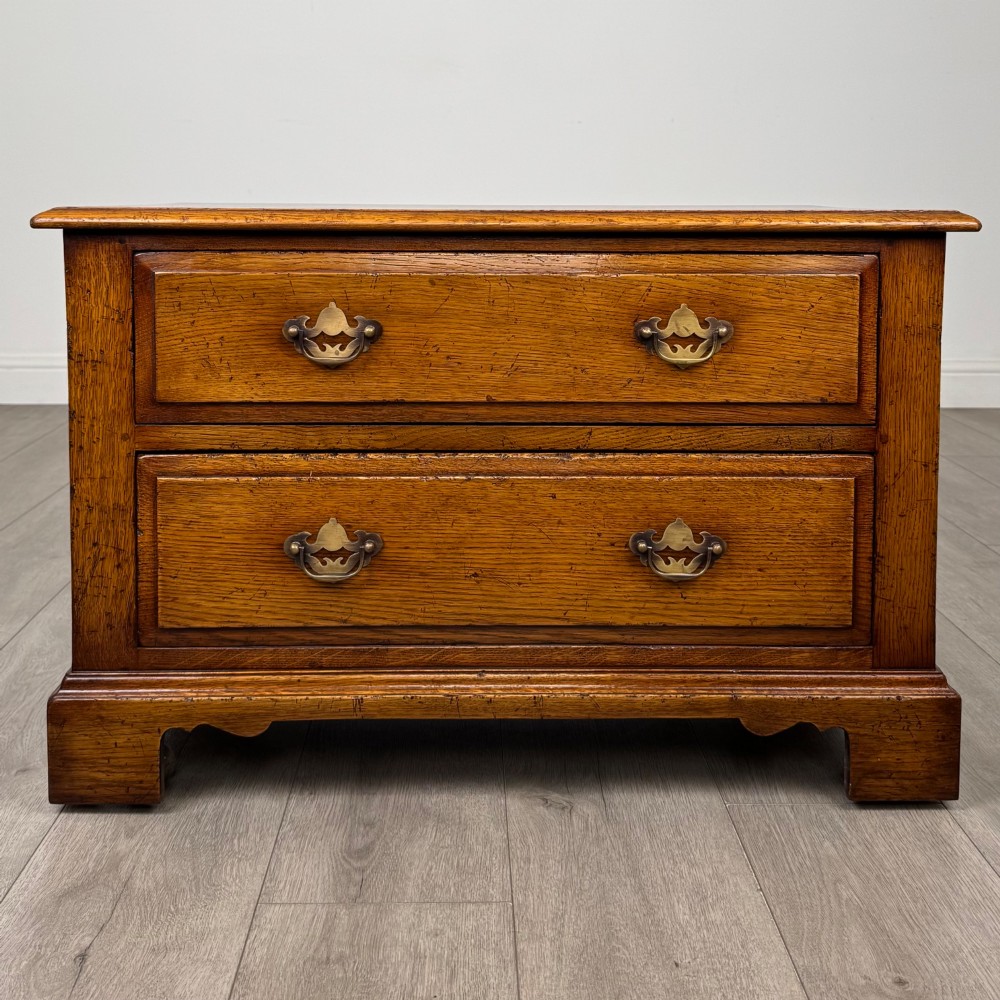 A low two-drawer oak chest featuring raised panel fronts, decorative brass drop handles, and bracket feet in a warm brown finish.