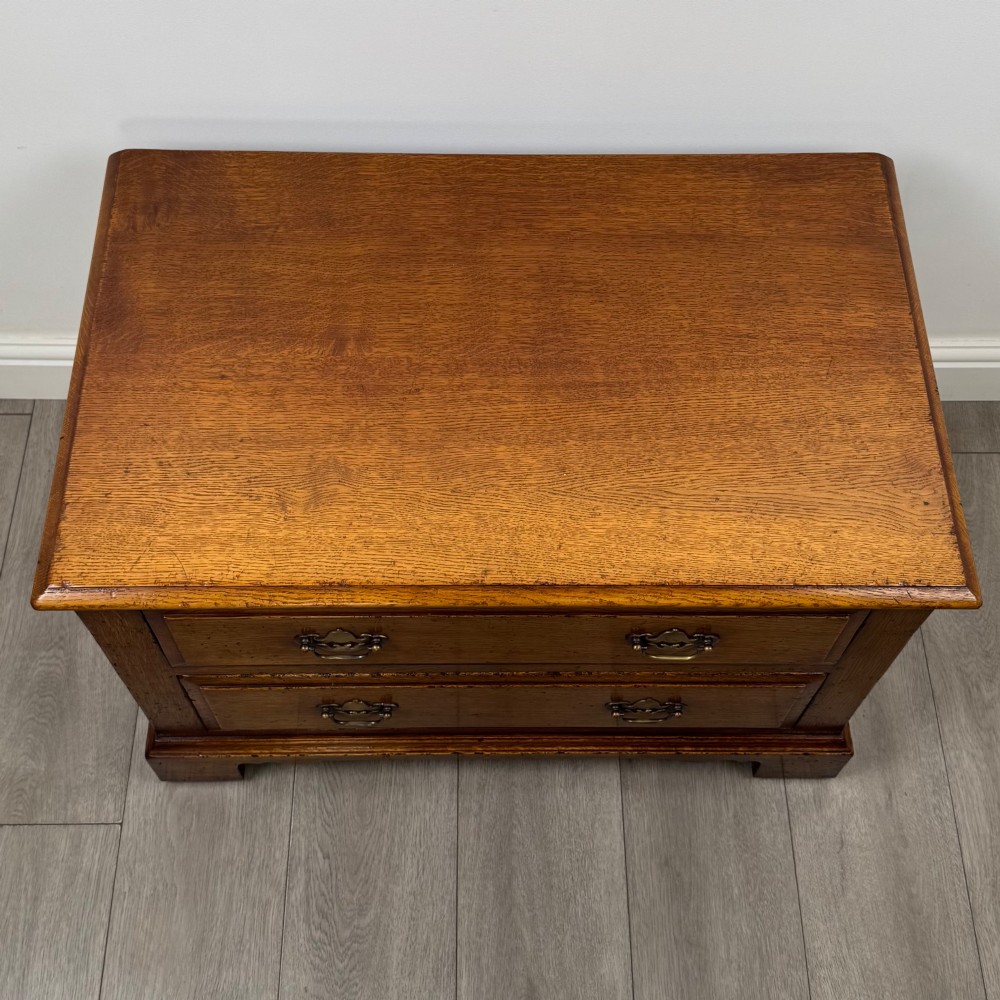 A low two-drawer oak chest featuring raised panel fronts, decorative brass drop handles, and bracket feet in a warm brown finish. View X