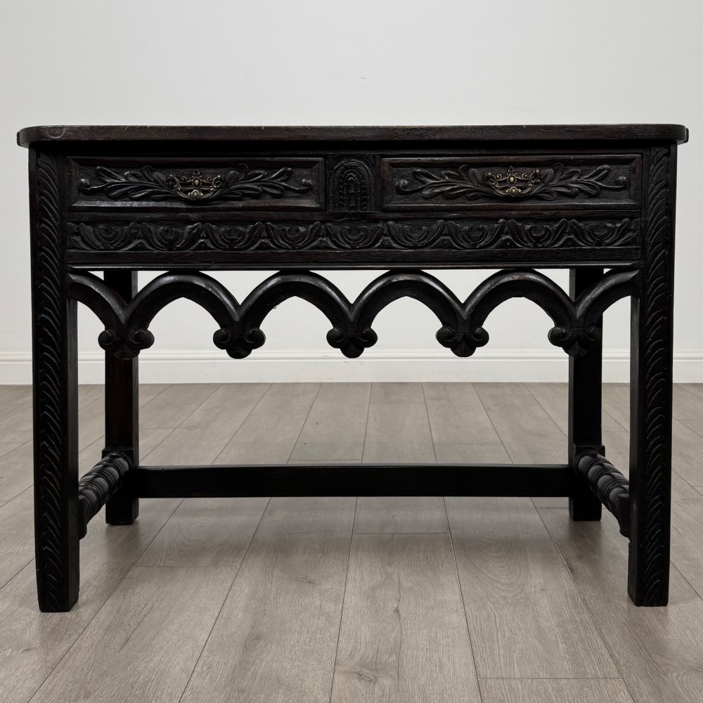 A dark oak 19th-century side table featuring floral relief carvings on two drawer fronts, a row of carved cusped arches along the lower apron, and turned side stretchers.
