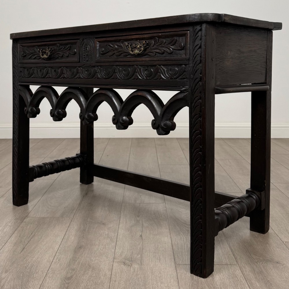 A dark oak 19th-century side table featuring floral relief carvings on two drawer fronts, a row of carved cusped arches along the lower apron, and turned side stretchers. View II