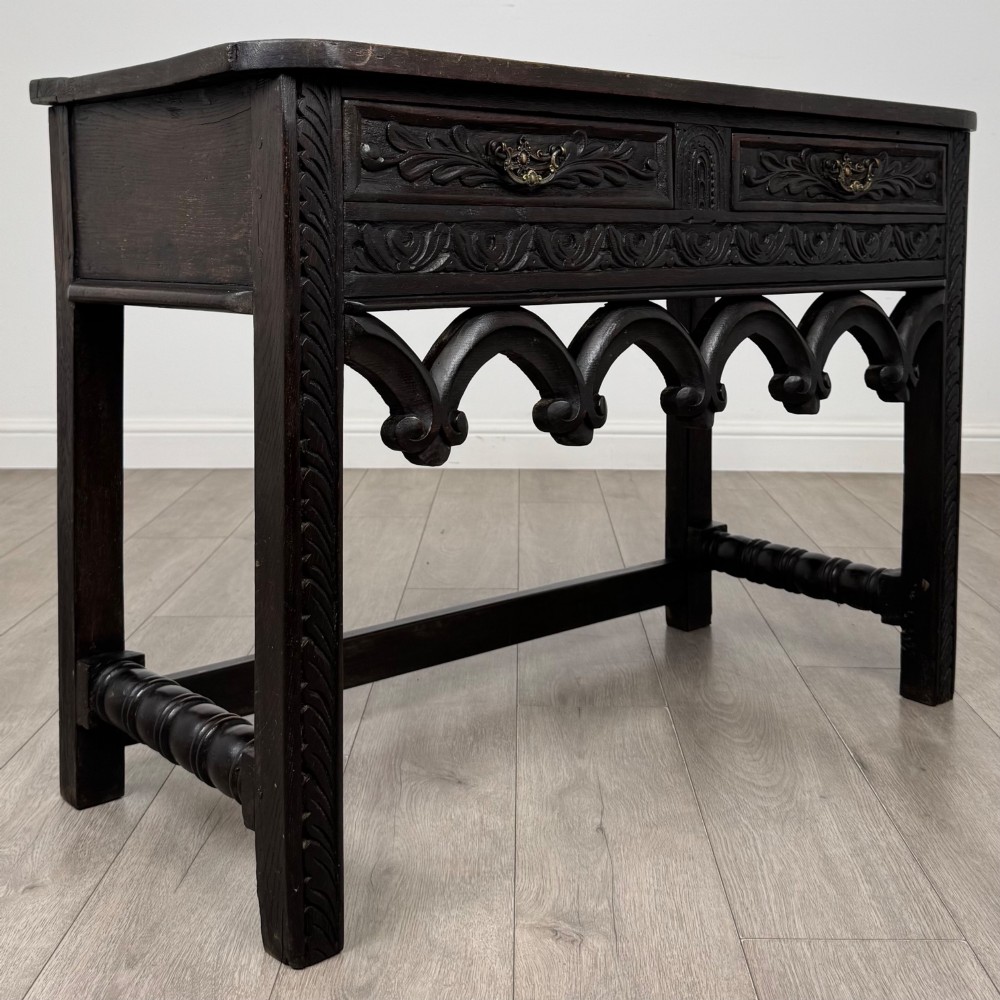 A dark oak 19th-century side table featuring floral relief carvings on two drawer fronts, a row of carved cusped arches along the lower apron, and turned side stretchers. View VIII