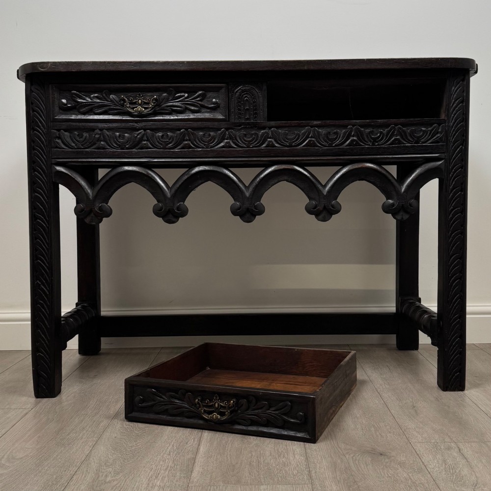 A dark oak 19th-century side table featuring floral relief carvings on two drawer fronts, a row of carved cusped arches along the lower apron, and turned side stretchers. View X