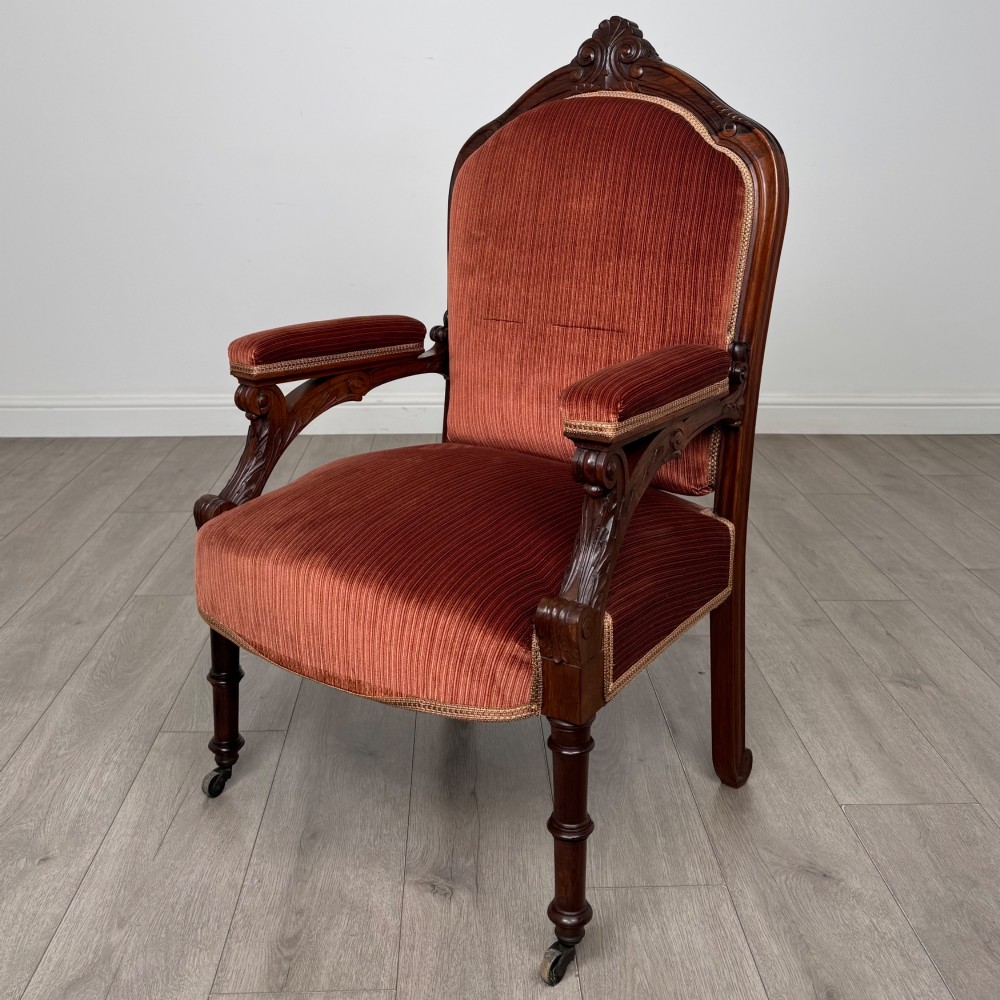 A Victorian rosewood armchair featuring a dark timber frame, carved scroll detailing, rust red velvet upholstery, and turned front legs on castors.