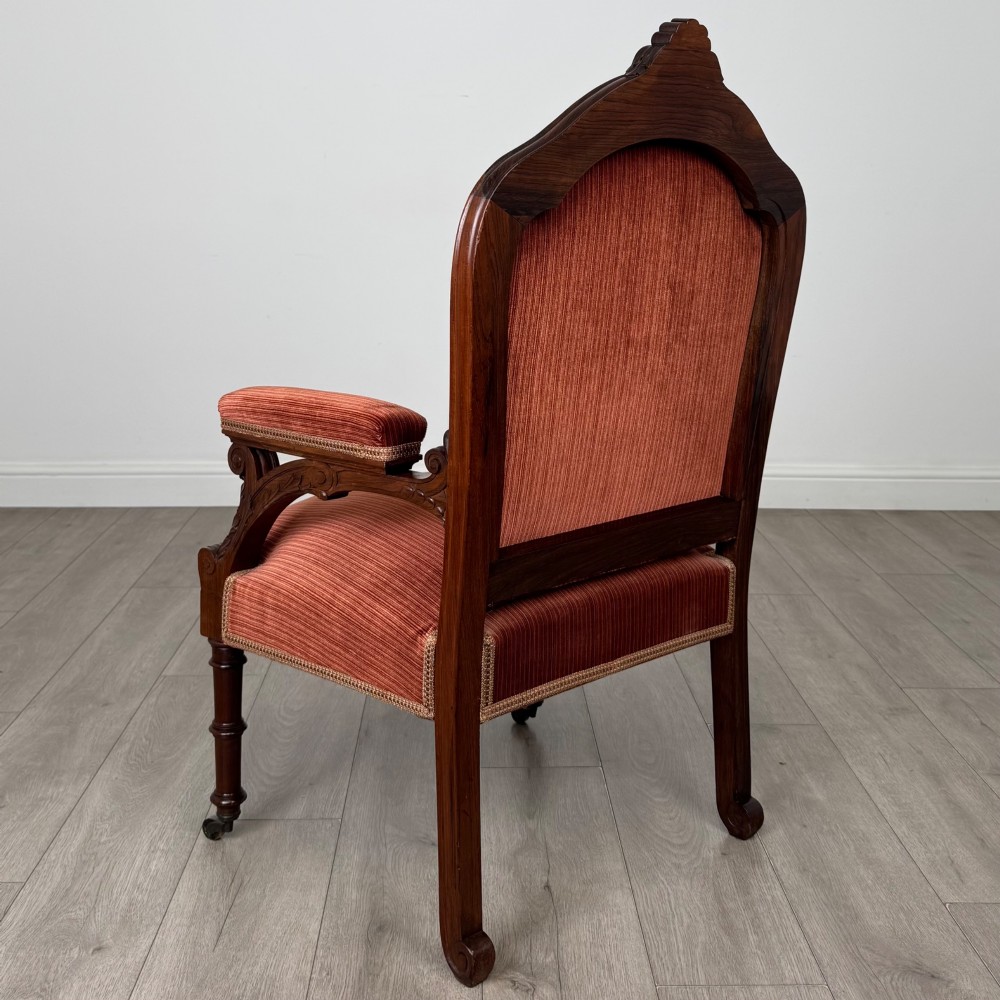 A Victorian rosewood armchair featuring a dark timber frame, carved scroll detailing, rust red velvet upholstery, and turned front legs on castors. View III