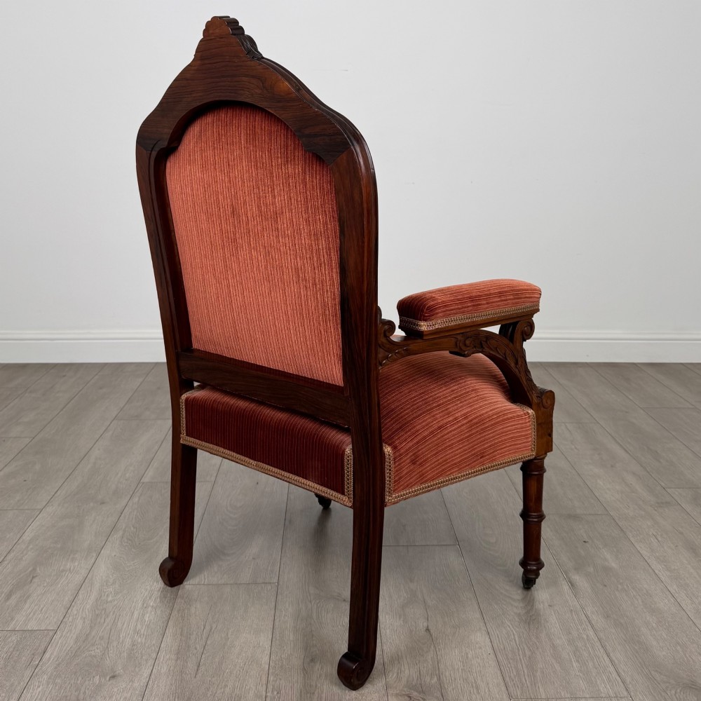 A Victorian rosewood armchair featuring a dark timber frame, carved scroll detailing, rust red velvet upholstery, and turned front legs on castors. View V