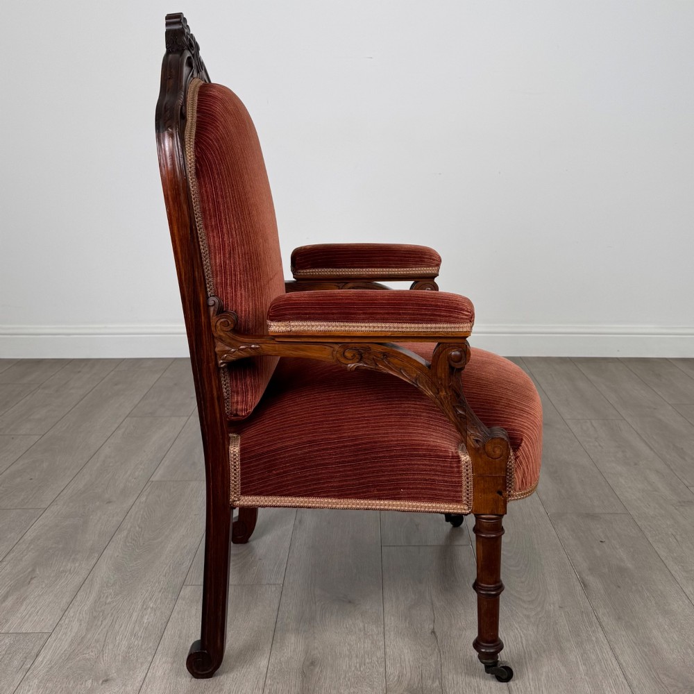 A Victorian rosewood armchair featuring a dark timber frame, carved scroll detailing, rust red velvet upholstery, and turned front legs on castors. View VI