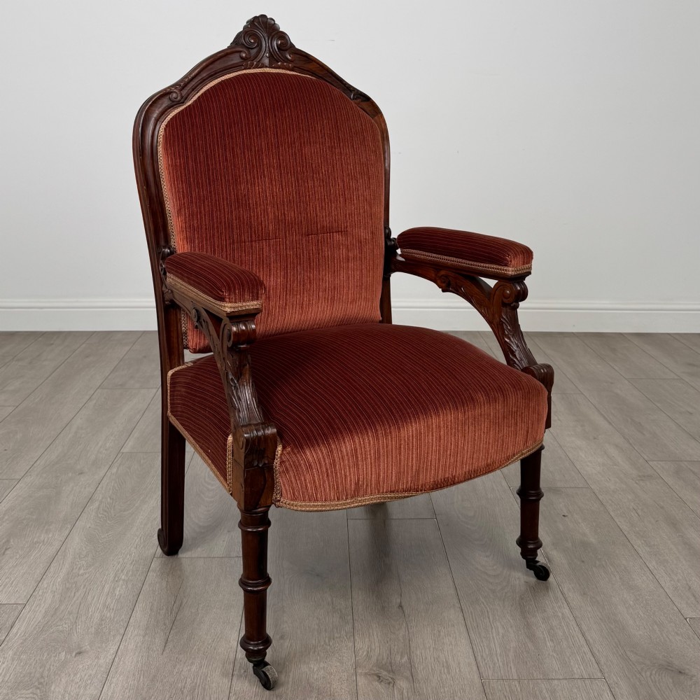 A Victorian rosewood armchair featuring a dark timber frame, carved scroll detailing, rust red velvet upholstery, and turned front legs on castors. View VII