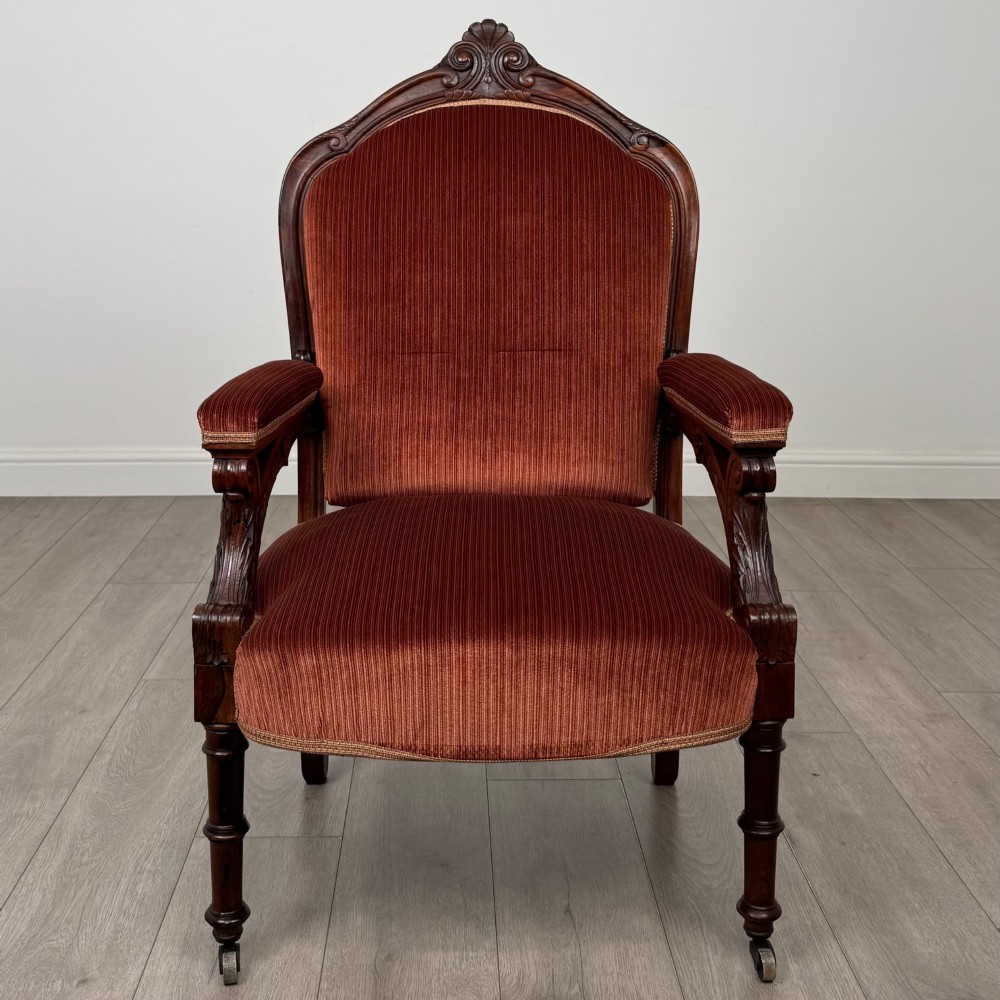A Victorian rosewood armchair featuring a dark timber frame, carved scroll detailing, rust red velvet upholstery, and turned front legs on castors. View VIII