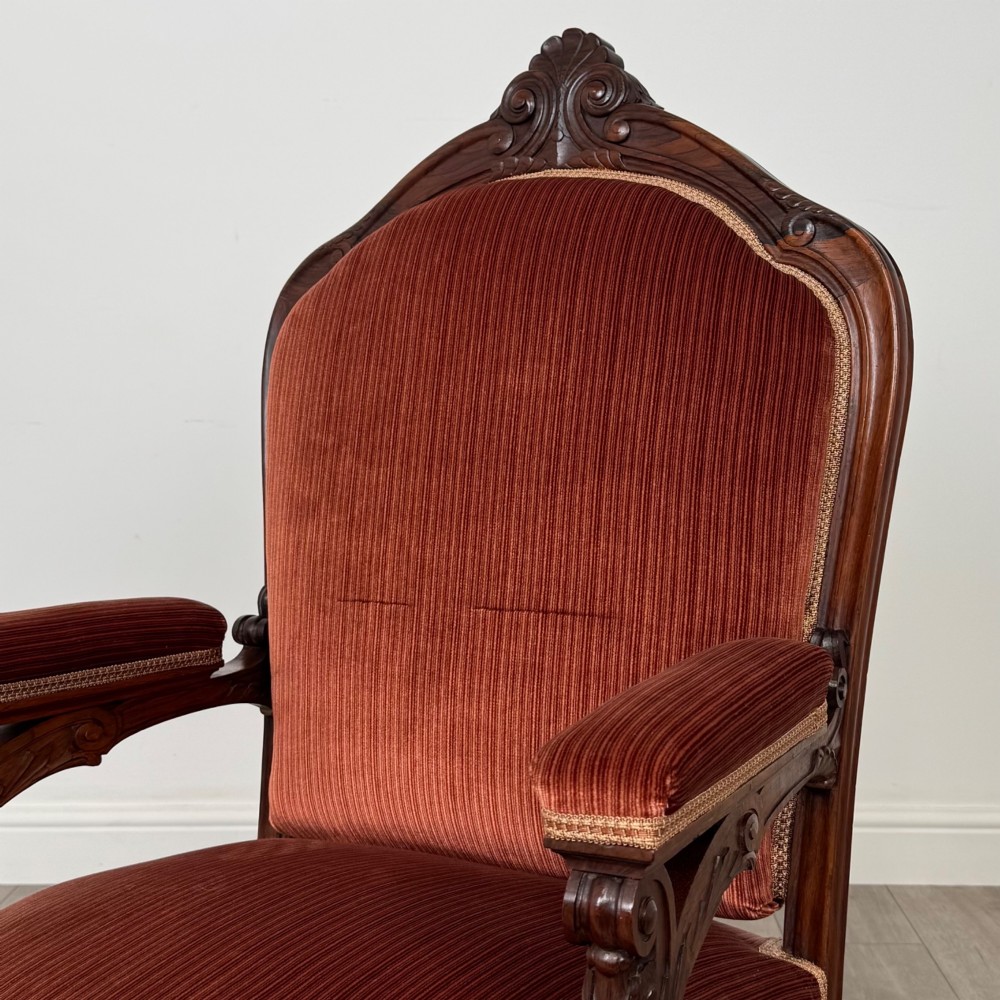 A Victorian rosewood armchair featuring a dark timber frame, carved scroll detailing, rust red velvet upholstery, and turned front legs on castors. View IX