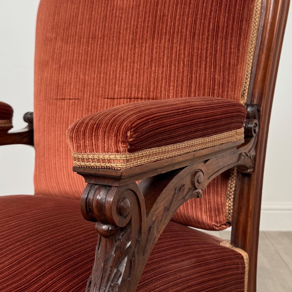A Victorian rosewood armchair featuring a dark timber frame, carved scroll detailing, rust red velvet upholstery, and turned front legs on castors. View X