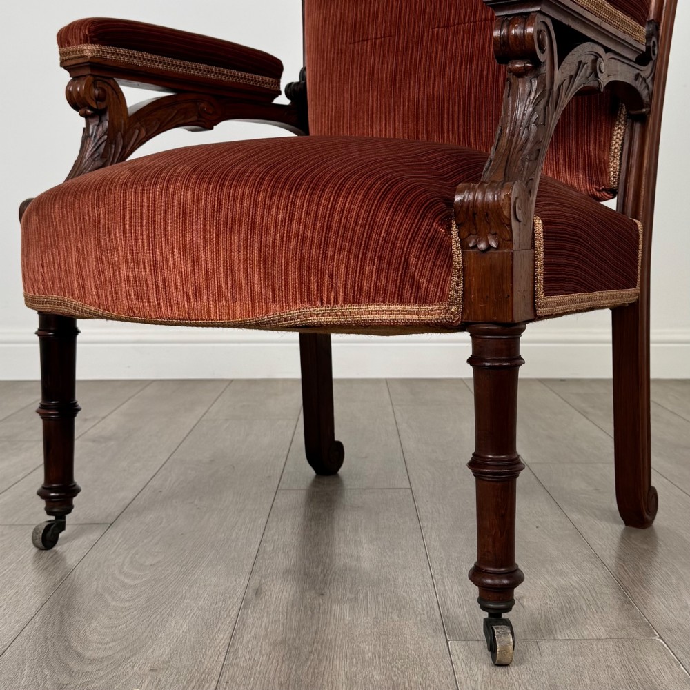 A Victorian rosewood armchair featuring a dark timber frame, carved scroll detailing, rust red velvet upholstery, and turned front legs on castors. View XI