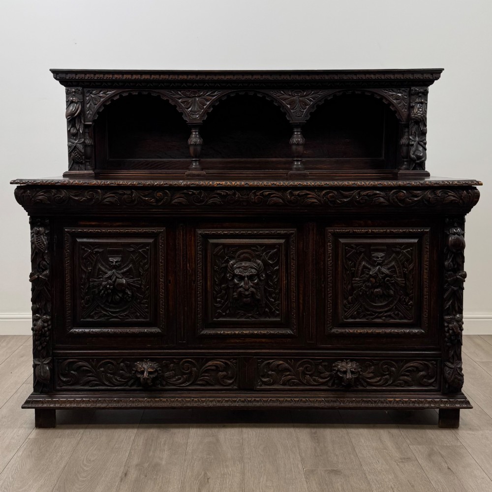 A dark oak 19th-century sideboard featuring deep relief carvings of Green Man mythical faces, foliate motifs, lion masks, and an arched upper gallery shelf.