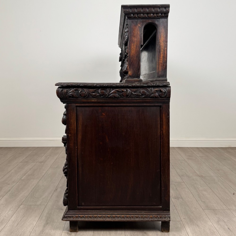 A dark oak 19th-century sideboard featuring deep relief carvings of Green Man mythical faces, foliate motifs, lion masks, and an arched upper gallery shelf. View IV