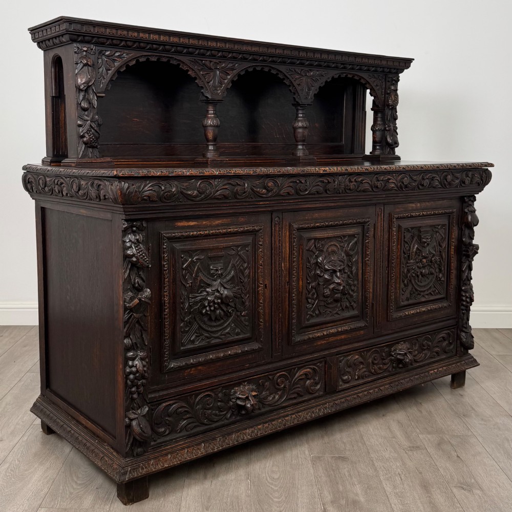 A dark oak 19th-century sideboard featuring deep relief carvings of Green Man mythical faces, foliate motifs, lion masks, and an arched upper gallery shelf. View IX