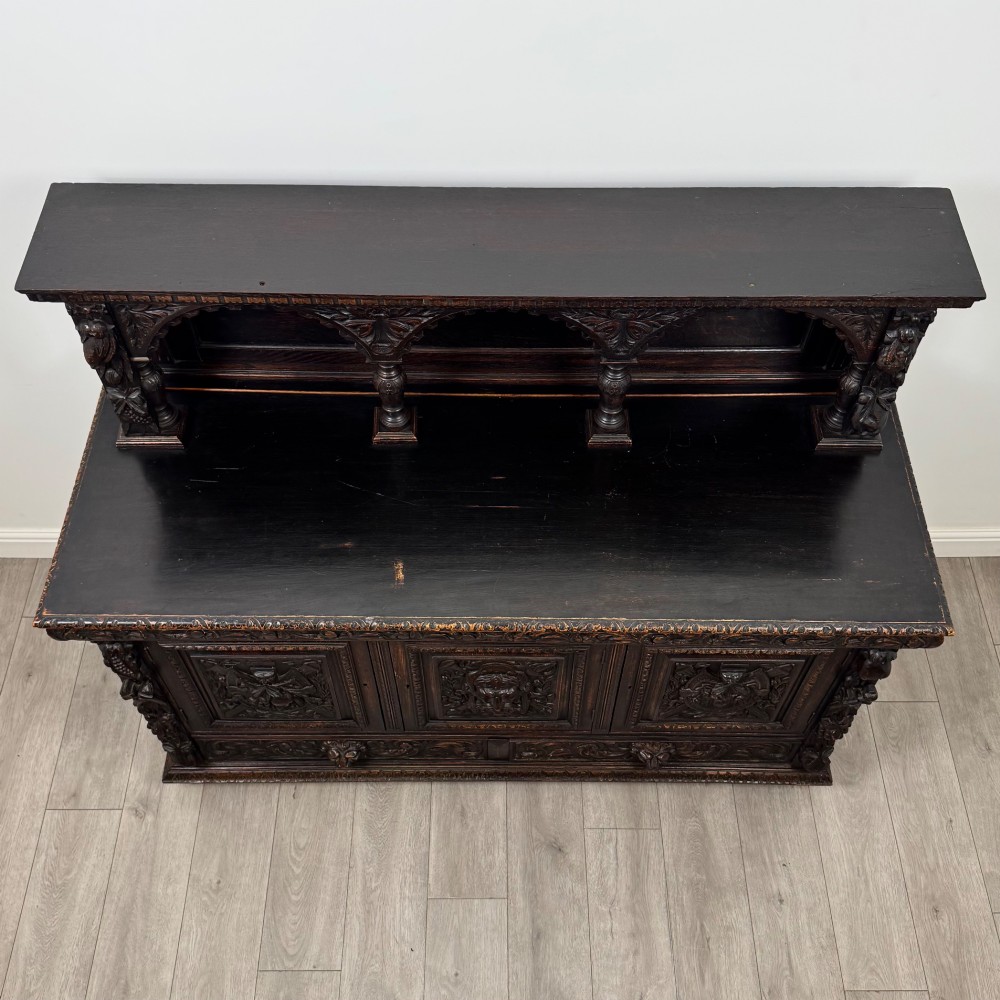 A dark oak 19th-century sideboard featuring deep relief carvings of Green Man mythical faces, foliate motifs, lion masks, and an arched upper gallery shelf. View X