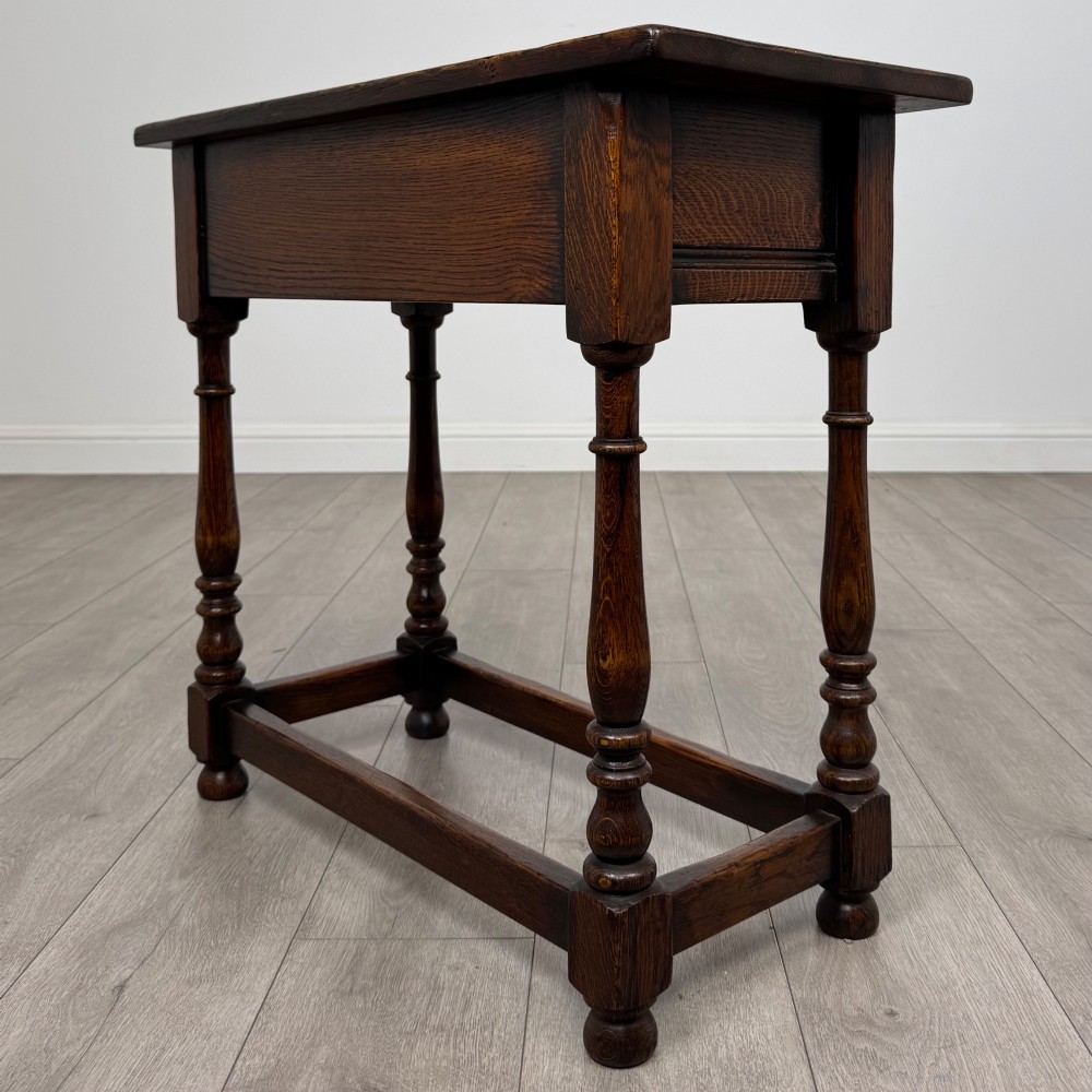 A dark oak side table featuring a single drawer with brass drop handles, turned legs, and a perimeter box stretcher. View VI
