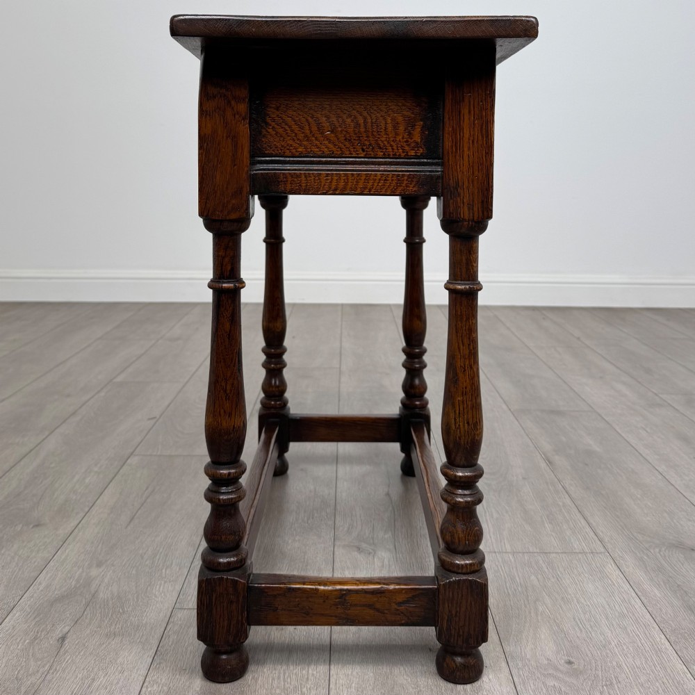 A dark oak side table featuring a single drawer with brass drop handles, turned legs, and a perimeter box stretcher. View VII