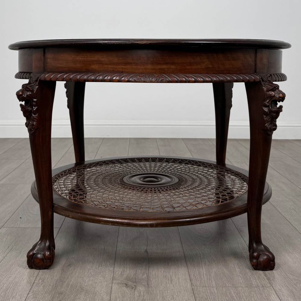 A circular dark mahogany coffee table featuring carved lion heads atop cabriole legs ending in claw and ball feet, with a woven cane lower shelf.