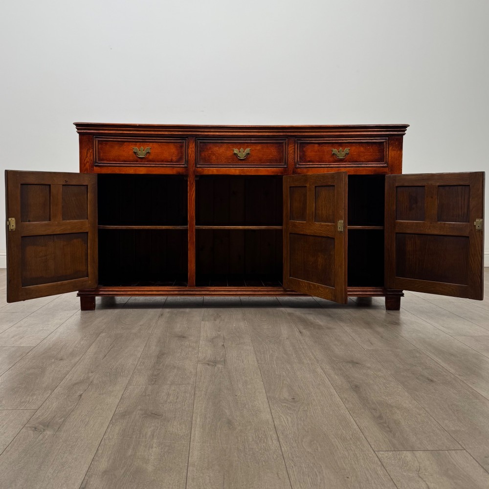 A twentieth-century warm oak sideboard featuring three upper drawers with brass handles above three cupboard doors decorated with geometric moulded panels and exterior brass H-hinges. View II