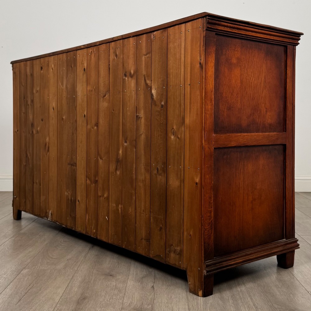 A twentieth-century warm oak sideboard featuring three upper drawers with brass handles above three cupboard doors decorated with geometric moulded panels and exterior brass H-hinges. View VII