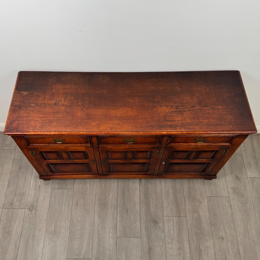 A twentieth-century warm oak sideboard featuring three upper drawers with brass handles above three cupboard doors decorated with geometric moulded panels and exterior brass H-hinges. View X