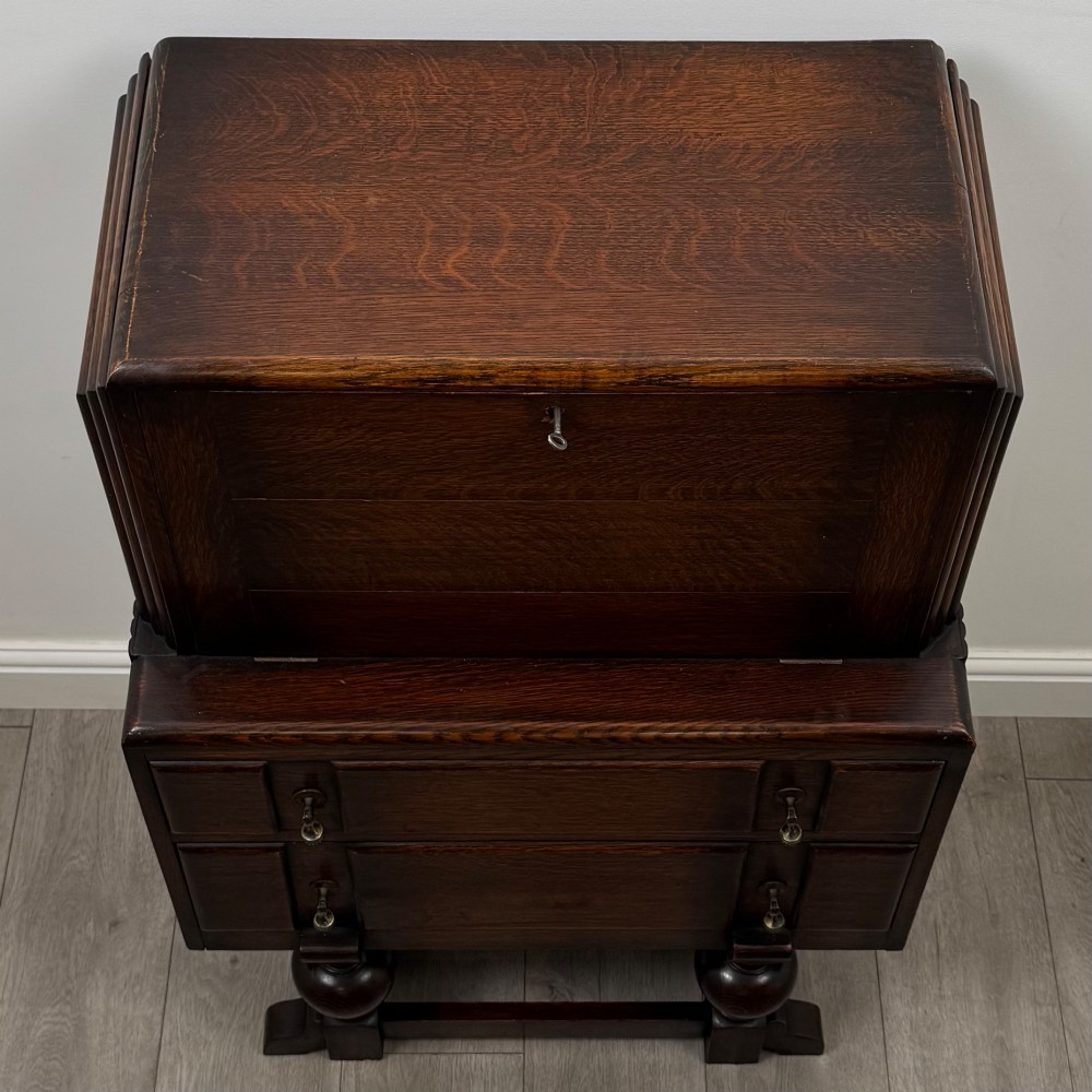 An early 20th-century dark oak writing bureau featuring a sloped fall-front lid above two drawers, supported on turned bulbous legs with stretchers. View X