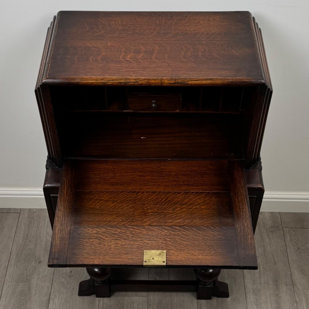 An early 20th-century dark oak writing bureau featuring a sloped fall-front lid above two drawers, supported on turned bulbous legs with stretchers. View XI