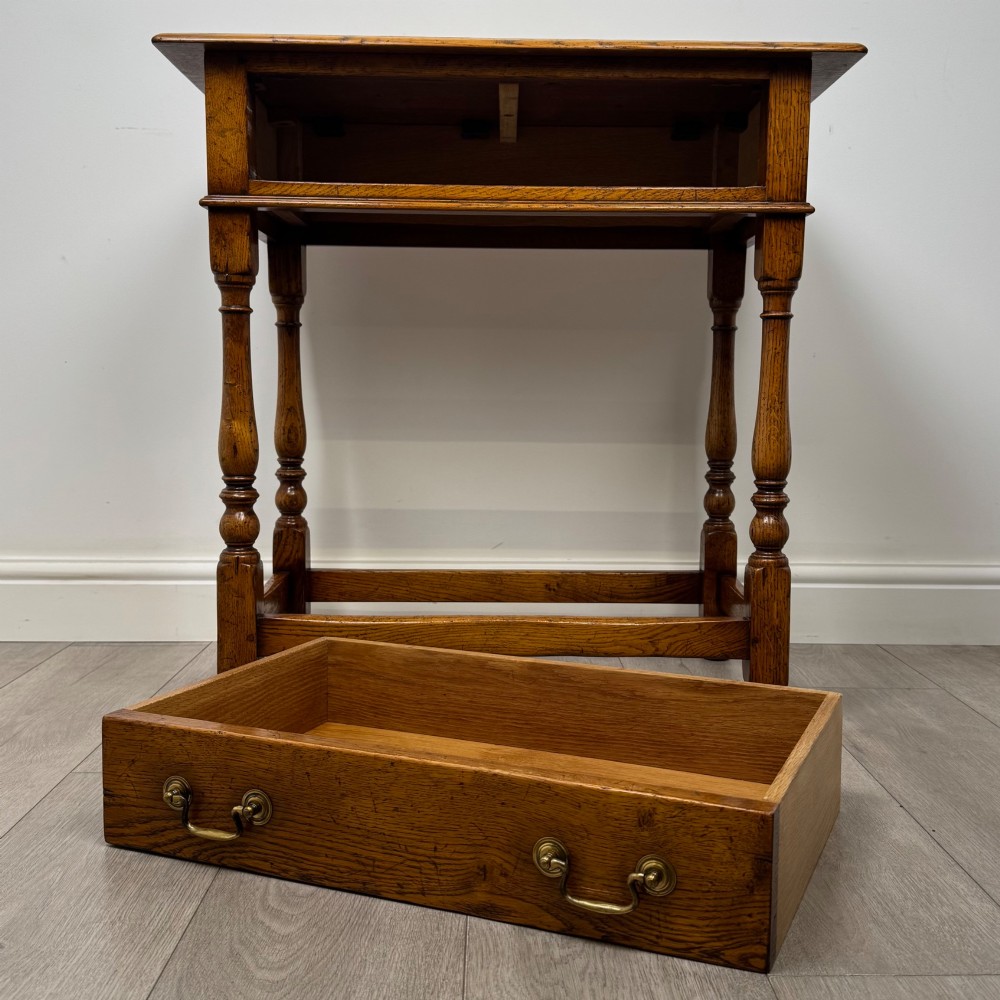 A golden oak side table featuring a single drawer with two brass handles, turned legs, and an lower stretcher base. View X