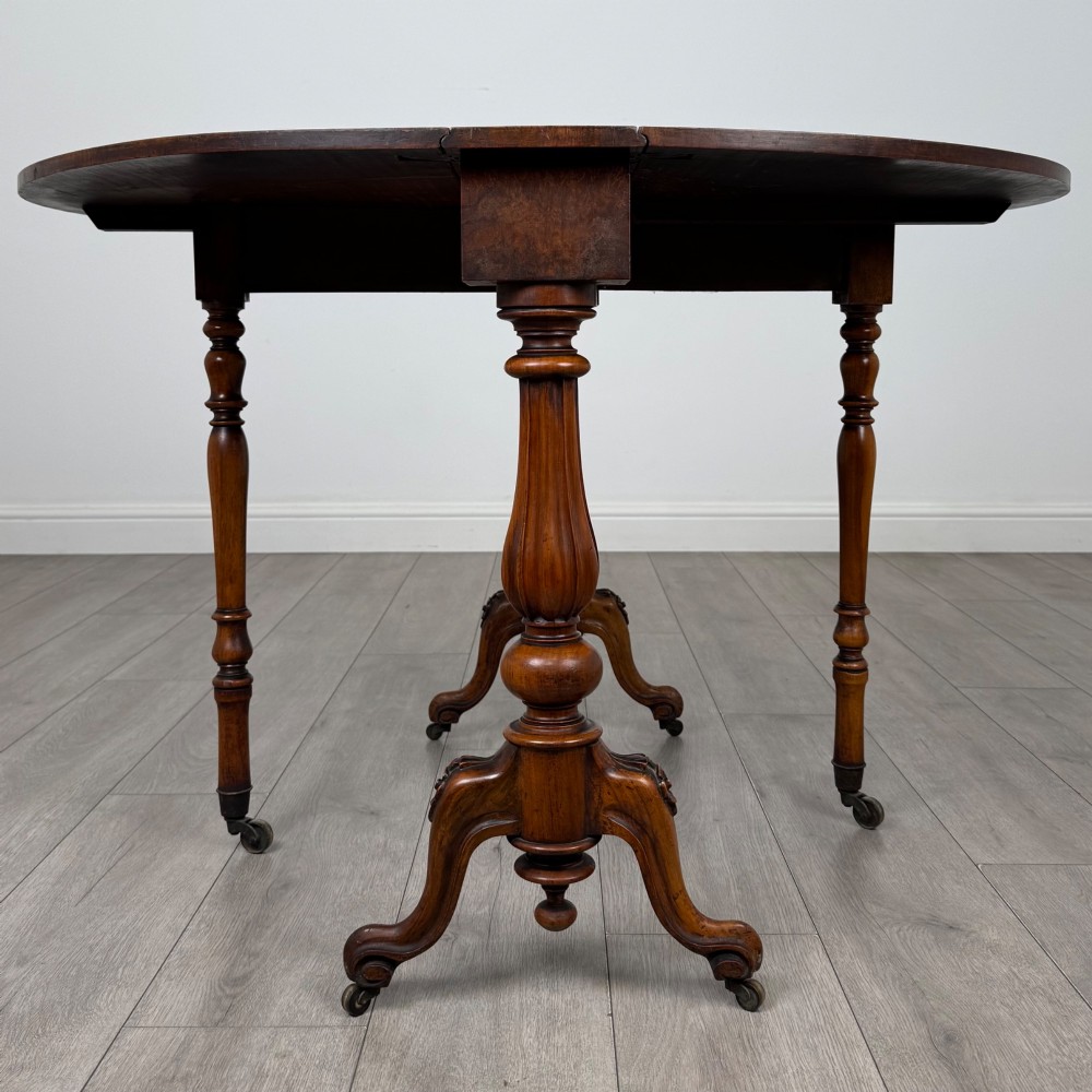 A 19th-century Victorian Sutherland table crafted from burr walnut featuring folded drop leaves, turned twin-column supports, and carved legs on brass castors. View V