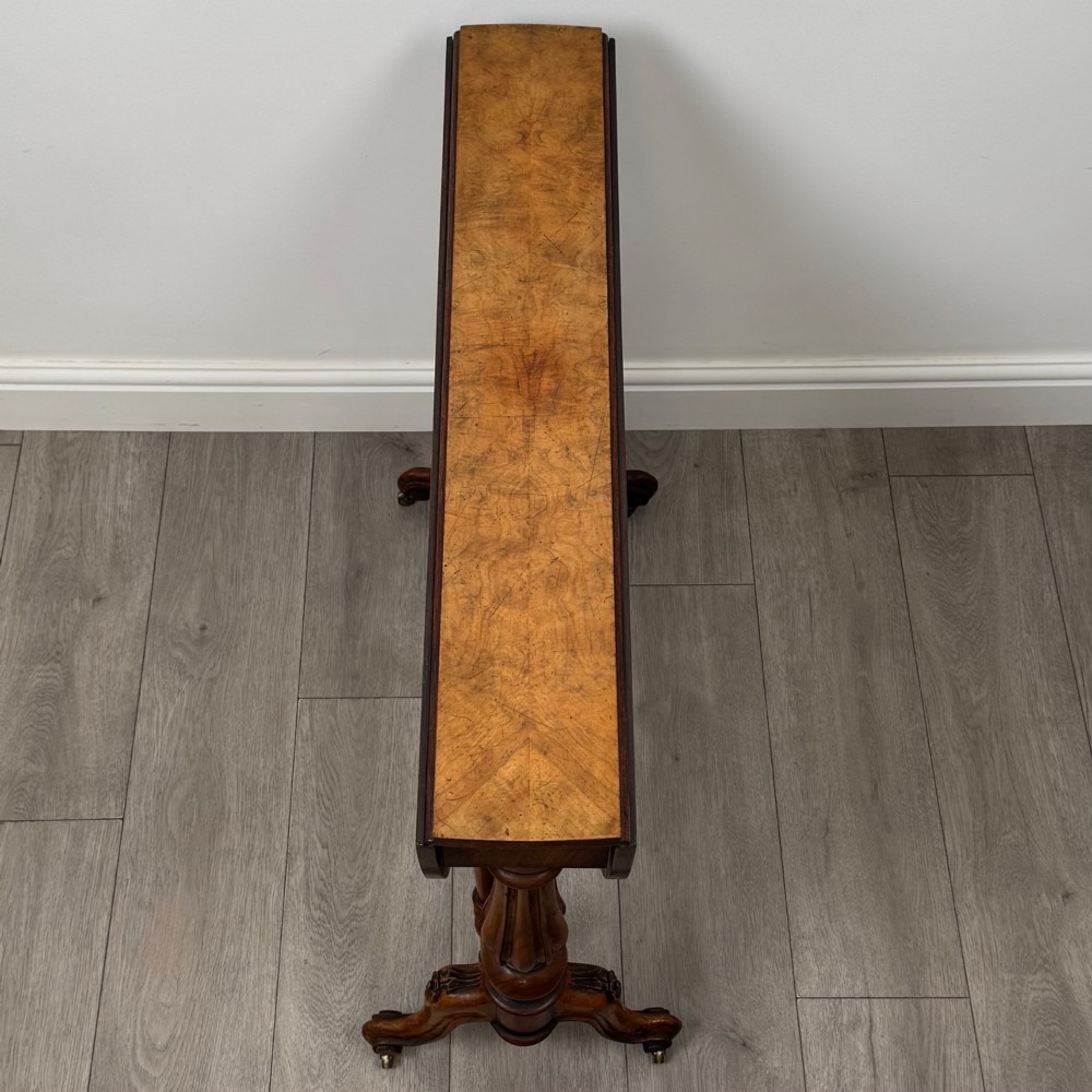 A 19th-century Victorian Sutherland table crafted from burr walnut featuring folded drop leaves, turned twin-column supports, and carved legs on brass castors. View X