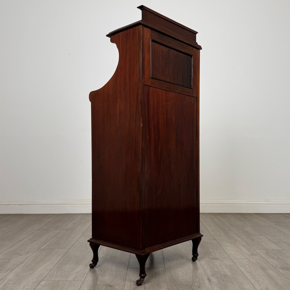 An early 20th-century mahogany music cabinet with an upper mirrored shelf, a middle glazed drawer compartment, a lower moulded panel door, and brass handles on castors. View V
