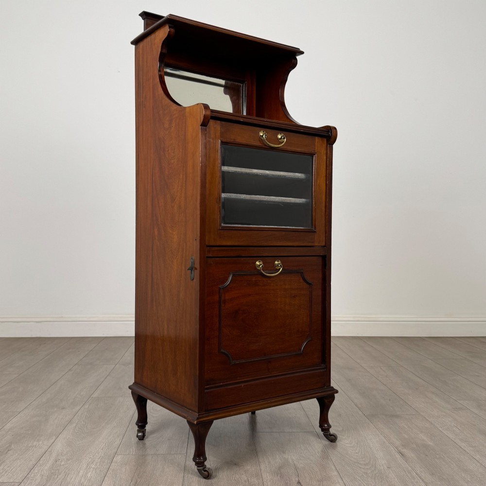 An early 20th-century mahogany music cabinet with an upper mirrored shelf, a middle glazed drawer compartment, a lower moulded panel door, and brass handles on castors. View IX