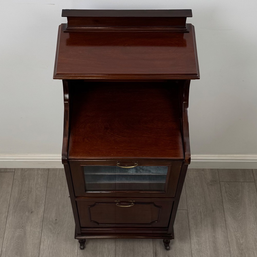An early 20th-century mahogany music cabinet with an upper mirrored shelf, a middle glazed drawer compartment, a lower moulded panel door, and brass handles on castors. View X