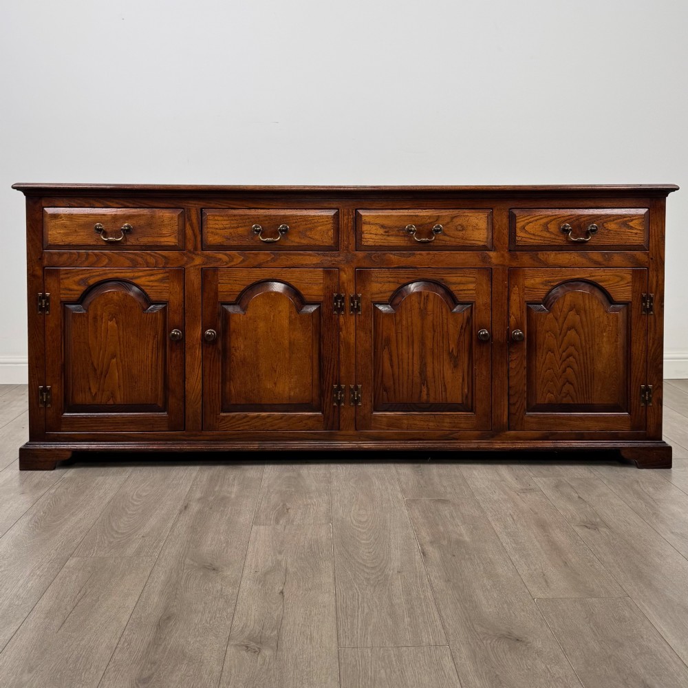 A large medium brown oak sideboard featuring four upper drawers with brass handles above four cupboard doors with arched wood panels.