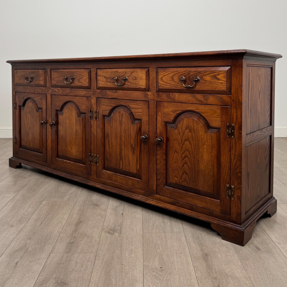 A large medium brown oak sideboard featuring four upper drawers with brass handles above four cupboard doors with arched wood panels. View III