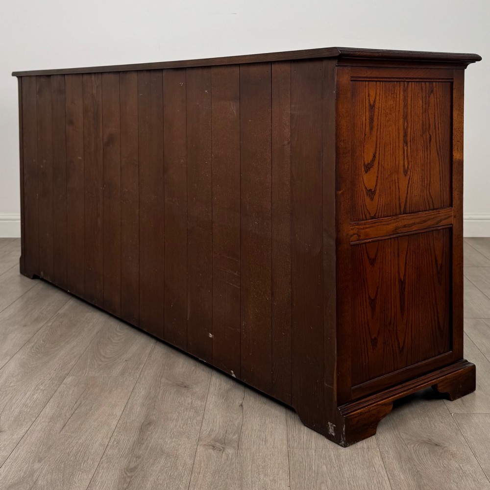 A large medium brown oak sideboard featuring four upper drawers with brass handles above four cupboard doors with arched wood panels. View VII