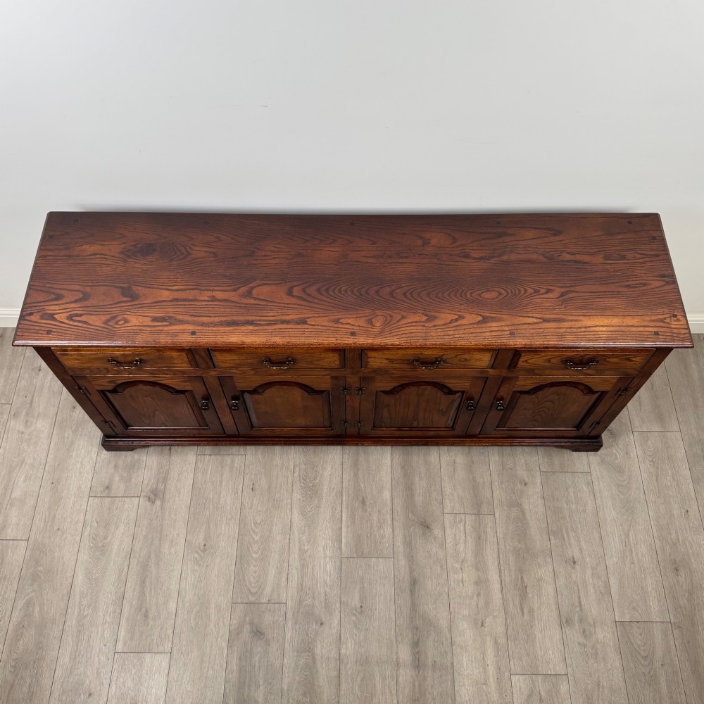 A large medium brown oak sideboard featuring four upper drawers with brass handles above four cupboard doors with arched wood panels. View X