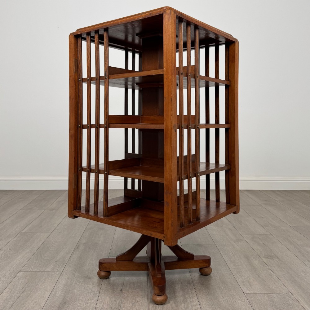 An Edwardian oak revolving bookcase with three tier shelves, open vertical slatted sides, and a central rotating column mounted on a quadruped base with castors. View II