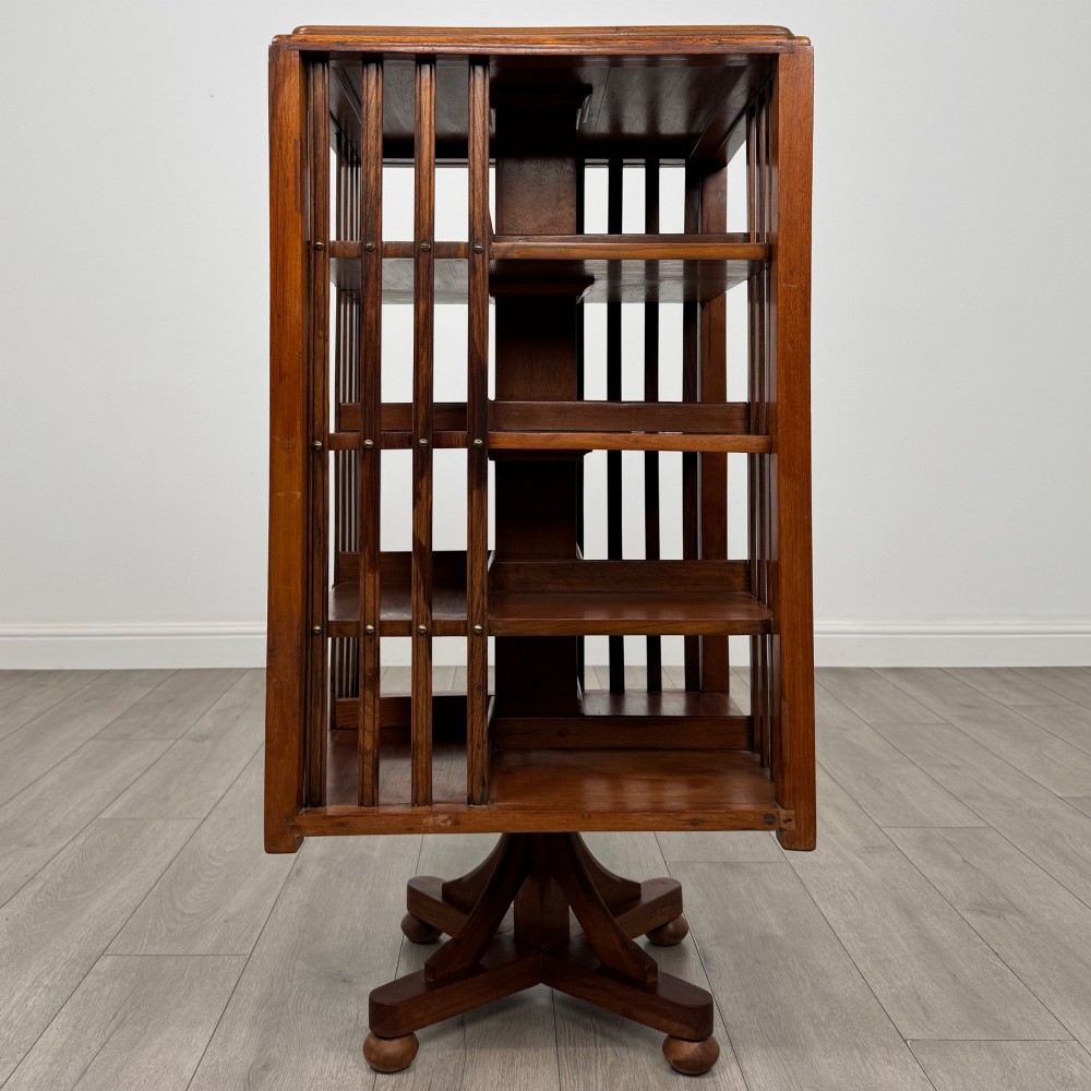 An Edwardian oak revolving bookcase with three tier shelves, open vertical slatted sides, and a central rotating column mounted on a quadruped base with castors. View V