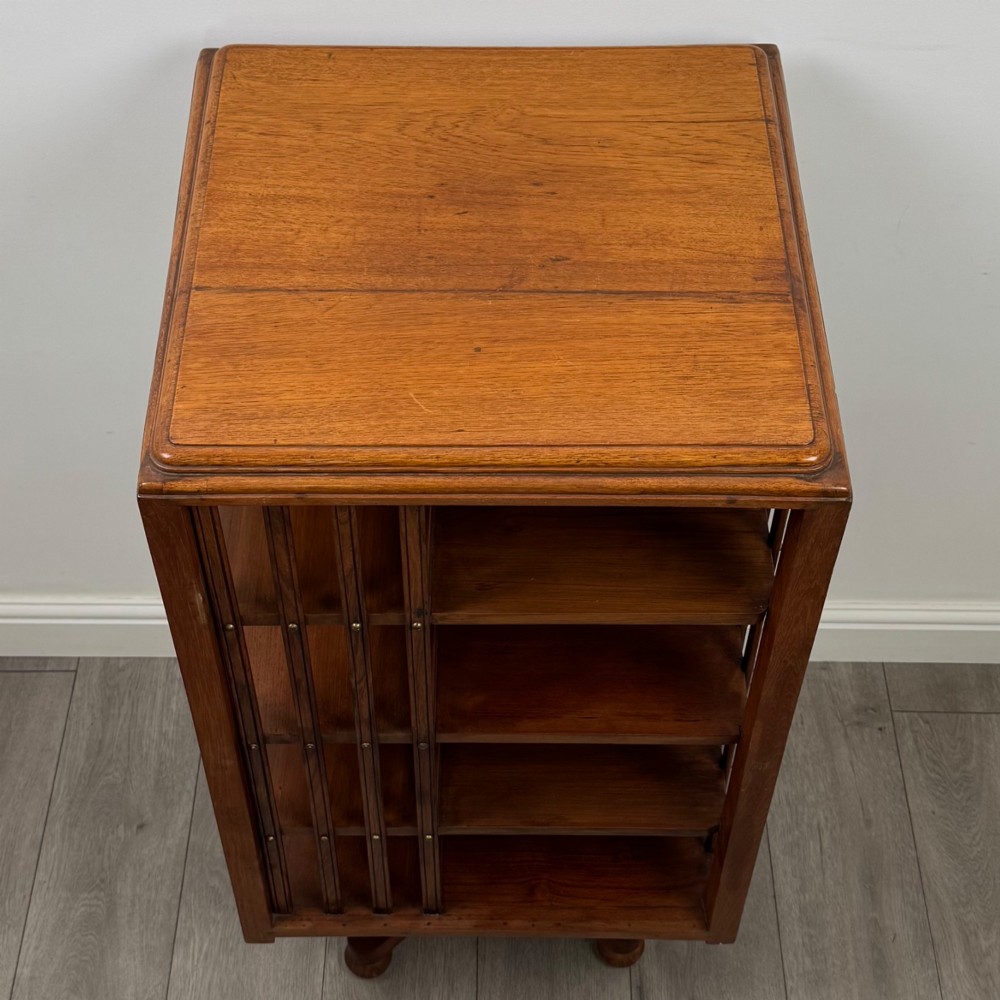 An Edwardian oak revolving bookcase with three tier shelves, open vertical slatted sides, and a central rotating column mounted on a quadruped base with castors. View VII