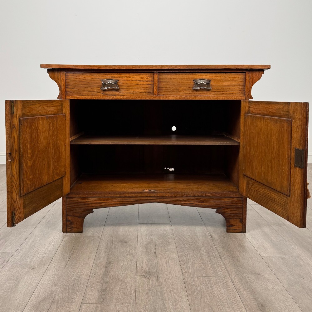 An Arts and Crafts golden oak sideboard featuring two top drawers, two lower cupboard doors, and stylized copper strap hinges. View II