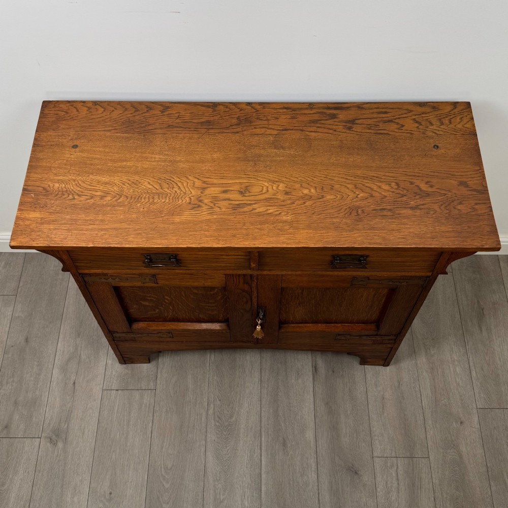 An Arts and Crafts golden oak sideboard featuring two top drawers, two lower cupboard doors, and stylized copper strap hinges. View X
