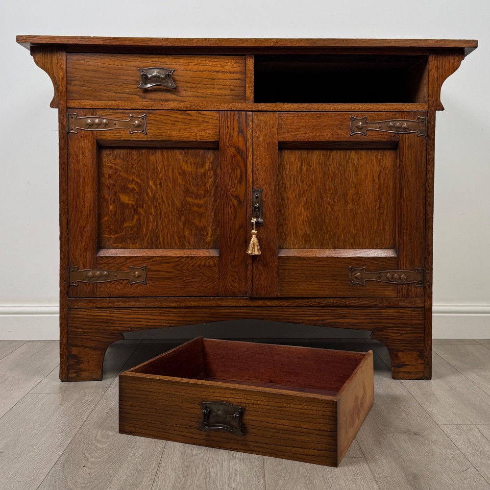 An Arts and Crafts golden oak sideboard featuring two top drawers, two lower cupboard doors, and stylized copper strap hinges. View XI