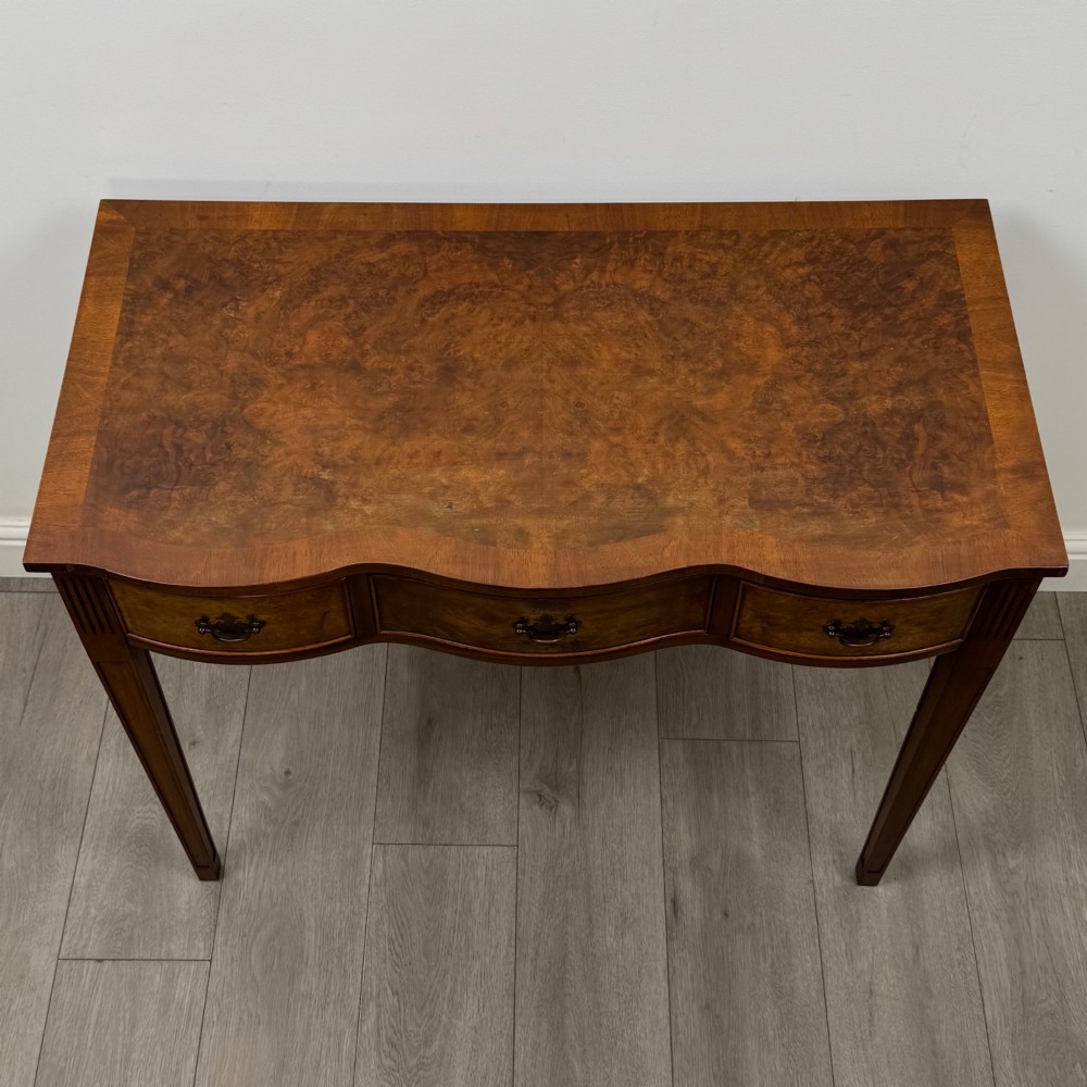A 20th-century burr walnut side table featuring a serpentine front, three drawer fronts with brass handles, and square tapered legs. View IX