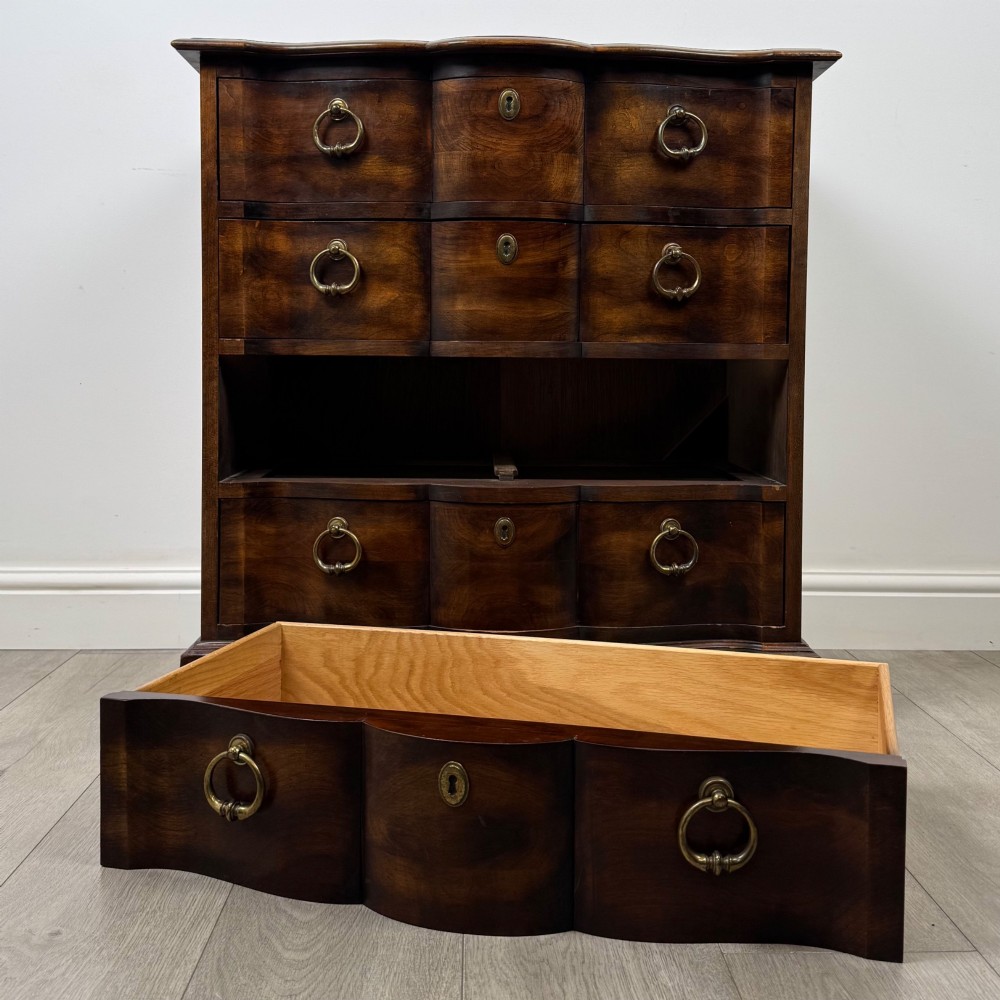 A 20th-century serpentine-front hardwood chest featuring four stacked drawers with brass drop ring handles and central brass escutcheons. View X