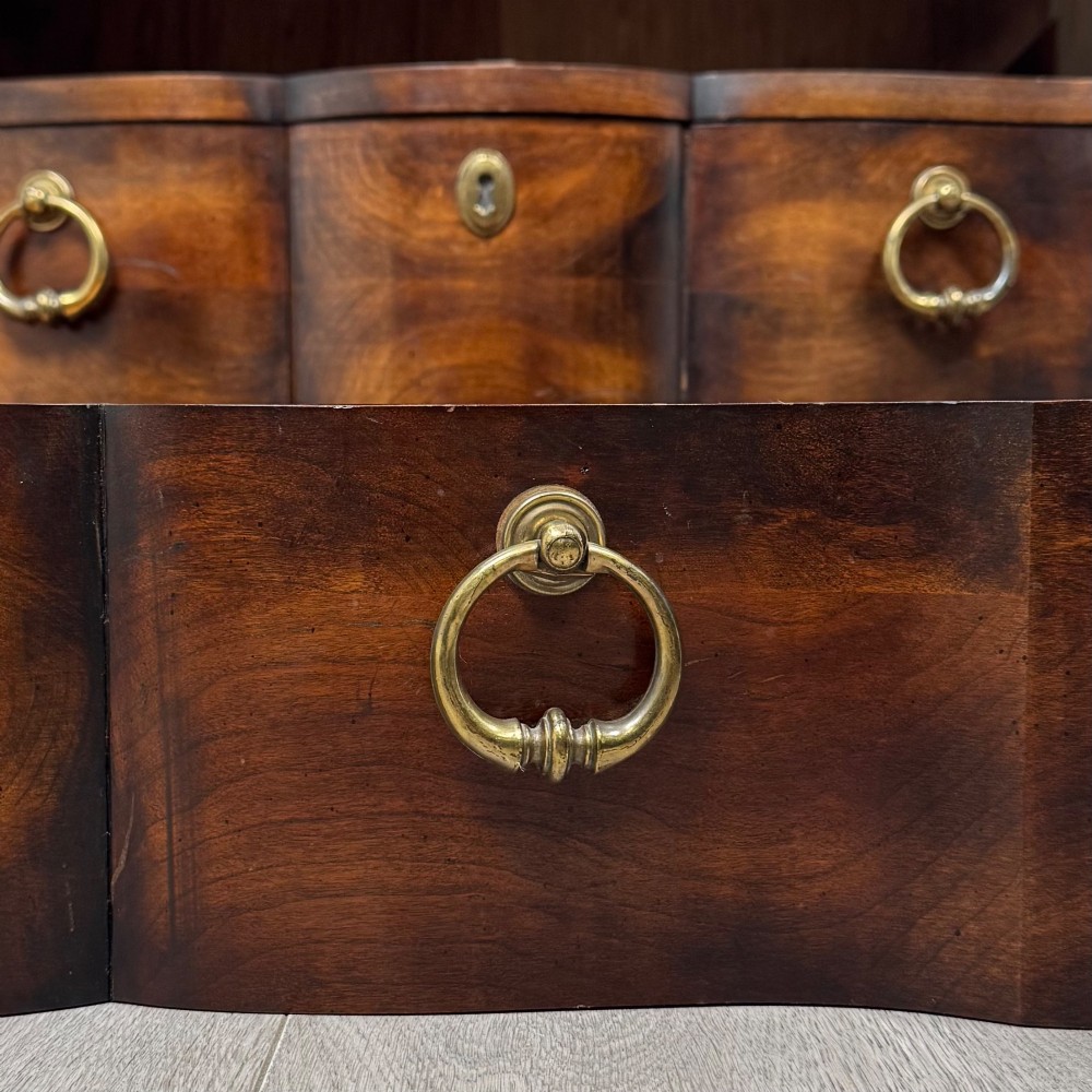 A 20th-century serpentine-front hardwood chest featuring four stacked drawers with brass drop ring handles and central brass escutcheons. View XI