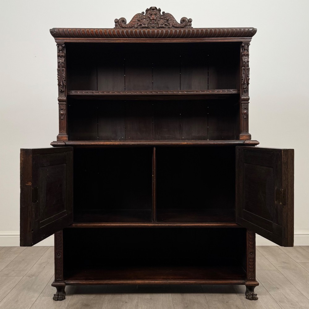 A 19th-century dark oak carved cupboard featuring an open top shelf, carved lion cresting, relief-carved panel doors with figures, and an open lower base. View II