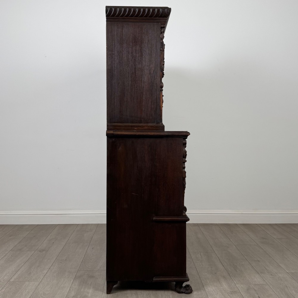 A 19th-century dark oak carved cupboard featuring an open top shelf, carved lion cresting, relief-carved panel doors with figures, and an open lower base. View VIII